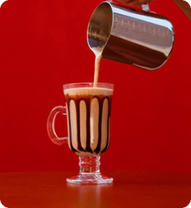 Imagem de uma taça cheia de cappuccino. Há uma pitcher derramando mais cappuccino na taça